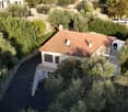 Aerial view of a two-story house with a red-tiled roof, surrounded by olive trees and greenery. The house features a balcony and a driveway leading to a parking area.