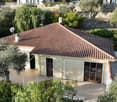 A single-story house with a terracotta-tiled roof, surrounded by olive trees and greenery. The house features large glass doors leading to a patio area.