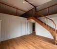 Empty room with wooden flooring, white walls, and a staircase leading to an upper level. The space includes a few doors and a visible section of a kitchen area in the background.