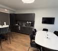 Modern, open-plan room with a kitchen area featuring dark cabinets and a coffee machine, two white tables with black chairs, and a flat-screen TV mounted on the wall.