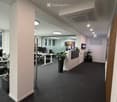 Modern office space with desks, chairs, computers, and plants. The area features a central column and large windows allowing natural light.
