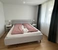 Bedroom with a double bed, nightstands, lamps, and curtains. The bed has a grey headboard and is made up with pink bedding.