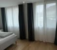 Minimalist bedroom with a single bed, nightstand, and lamp. Large windows with dark curtains allow natural light to fill the space.