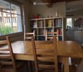 Bright room with large windows, wooden dining table with chairs, bookshelf filled with books, and a small kitchen area with a dishwasher.