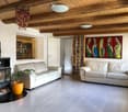 Bright living room with exposed wooden beams, two white sofas, a fireplace, a potted plant, and a colorful abstract painting on the wall.