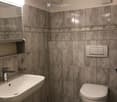 Modern bathroom with grey tiled walls and floor, white sink with chrome faucet, toilet, mirror, and light switch.