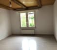 Empty room with wooden flooring, white walls, large window with double panes, radiator below window, exposed wooden ceiling beams, and a single light fixture.