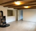 Empty room with wooden flooring, exposed wooden ceiling beams, and a black wood-burning stove with a chimney. There is a window and an open doorway leading to another room.