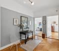 Modern hallway with wooden flooring, grey walls, a small black table with a vase and picture frames, white wire chairs, and an open doorway leading to a living area with a sofa and additional decor.