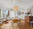 Bright dining room with wooden herringbone flooring, large windows, a round dining table with six chairs, a wooden cabinet, a round mirror, and decorative lamps.