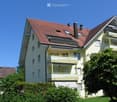 Two-story house with red-tiled roof, multiple windows, balconies, and surrounding greenery.