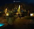 Nighttime outdoor area with a swimming pool, stone-paved ground, large thatched-roof structure, tables and chairs, decorative lanterns, and potted plants.