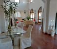Elegant living and dining area with wooden flooring, white walls, arched doorways, and large windows. Features a glass dining table with four chairs, a cream-colored sofa, a decorative vase, and a tall plant in a pot.