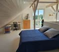 Modern attic bedroom with slanted wooden ceilings, large windows, a bed with blue bedding, a desk with a chair, a cabinet, and a small shelf with colorful storage bins.