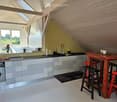 Modern kitchen with grey cabinets, white countertops, and a red dining table with stools. Features a dishwasher, sink, and a view of the outdoors through large windows.
