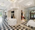 Elegant hallway with checkered black and white flooring, white walls, ornate chandeliers, and several round tables with white tablecloths, surrounded by chairs and decorative flower arrangements.