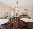 Elegant dining area with round tables, chairs, ornate chandelier, large windows with curtains, and a patterned carpet.