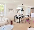 Bright, open living and dining area with light wood flooring, a beige sofa, a glass coffee table, a modern dining table with chairs and a wooden bench, and decorative vases.