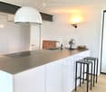 Modern kitchen with white cabinets, a central island, and a sleek countertop. Features include a hanging pendant light, two bar stools, a small fan, and a decorative bowl on the island.