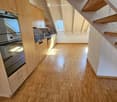 Modern kitchen with light wooden cabinets, stainless steel appliances, and a polished wooden floor. The space is bright with natural light from skylights.
