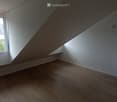 Empty attic room with wooden flooring, white walls, and large windows offering a view of the outside. The ceiling has a sloped design typical of attic spaces.