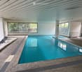Indoor swimming pool area with large windows, tiled pool, lounge chairs, and a modern ceiling design.