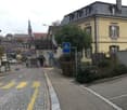 A quiet street lined with multi-story residential buildings, featuring classic European architecture with pitched roofs and chimneys. The road is paved with cobblestones and marked with yellow pedestrian crossing lines. A small garden area is visible in f