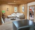 A cozy living room with wooden walls and ceiling, a sectional sofa, coffee table, and various chairs and stools. Artwork and a desk are also present.