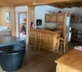 A cozy, wooden-paneled room with a central bar, bar stools, a leather chair, a sofa, and a small kitchen area with cabinets and a refrigerator.