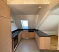 Modern kitchen with light wooden cabinets, black countertops, and built-in appliances including an oven and dishwasher. Features a skylight window and wooden flooring.