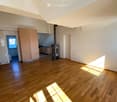 Empty room with wooden flooring, white walls, and a fireplace in the corner. Sunlight streams in from a window on the right side.