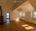 Bright, empty attic room with wooden flooring, white walls, and large windows offering a scenic mountain view. Features a fireplace and a sliding glass door leading to a balcony.