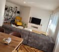 Modern living room with grey sectional sofa, wooden coffee table, yellow armchair, bookshelf, TV stand, and decorative items.