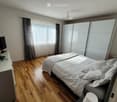 Modern bedroom with wooden flooring, large bed with white bedding, wardrobe, and large window with curtains.