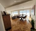 Modern living and dining area with herringbone wooden flooring, large windows with sheer curtains, a grey sofa with ottoman, a wooden dining table with four chairs, and a potted plant near the window.
