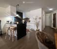 Modern kitchen with white cabinets, black island, and wooden flooring. Features two stools at the island, a fur throw on a chair, and decorative candles.