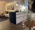 Modern living room with a sectional sofa, coffee table, floor lamp, and decorative items. Features a sleek black and white bar area with a faucet and candle, along with framed artwork and a tall plant in the corner.