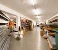 A spacious, well-lit warehouse or storage area with exposed white ceiling beams, metal shelving units, and various construction materials and tools. The floor is concrete, and the space is organized with pallets, boxes, and stacked items.