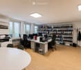 Office space with wooden flooring, large windows, desks, chairs, computers, printer, bookshelf, and coat rack.