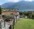 A scenic garden area with a stone wall, wooden railing, and a round stone table, overlooking a lake and mountains.