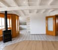 Bright, open living space with wooden floors, white walls, and a black wood-burning stove. Features include wooden doors, built-in wooden shelves, and a small kitchen area with white cabinets.