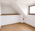 Empty attic room with wooden flooring, white walls, and exposed wooden beams on the ceiling. Features a single window with wooden frame and a built-in wooden shelf.