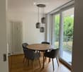 Dining area with a round wooden table, four grey chairs, pendant lights, and large windows overlooking a balcony.