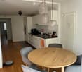 Modern kitchen and dining area with light wood flooring, white cabinets, a round wooden dining table with four grey upholstered chairs, and pendant lights above the table. The kitchen features a black countertop with a sink, coffee maker, and other small