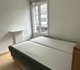 A small bedroom with a single bed, wooden flooring, and a window with blinds. The bed has a white mattress and dark grey bedding.