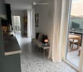 Modern open-plan living and dining area with a sofa, small table, and a kitchen counter. Large windows lead to a balcony with a small table and chairs.