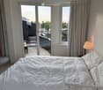 Bright bedroom with a white bed, nightstand, and lamp. Large sliding glass doors open to a balcony with outdoor seating.