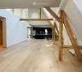 Empty hallway with wooden flooring, light blue walls, and exposed wooden beams. The hallway leads to a kitchen area with dark cabinets and stainless steel appliances visible in the background.