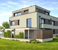 Modern two-story house with a raised ground floor, large windows, and a balcony on the upper level. The house features a combination of light and dark tones, surrounded by a well-maintained lawn and greenery.