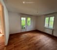 Empty room with wooden flooring, white walls, and large windows allowing natural light. Features a radiator and a door leading to another space.